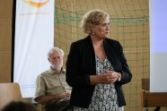 Frau steht in einem Raum, dahinter sitzt ein Mann