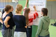 Jugendliche stehen vor einer Pinnwand, eine Person schreibt etwas darauf
